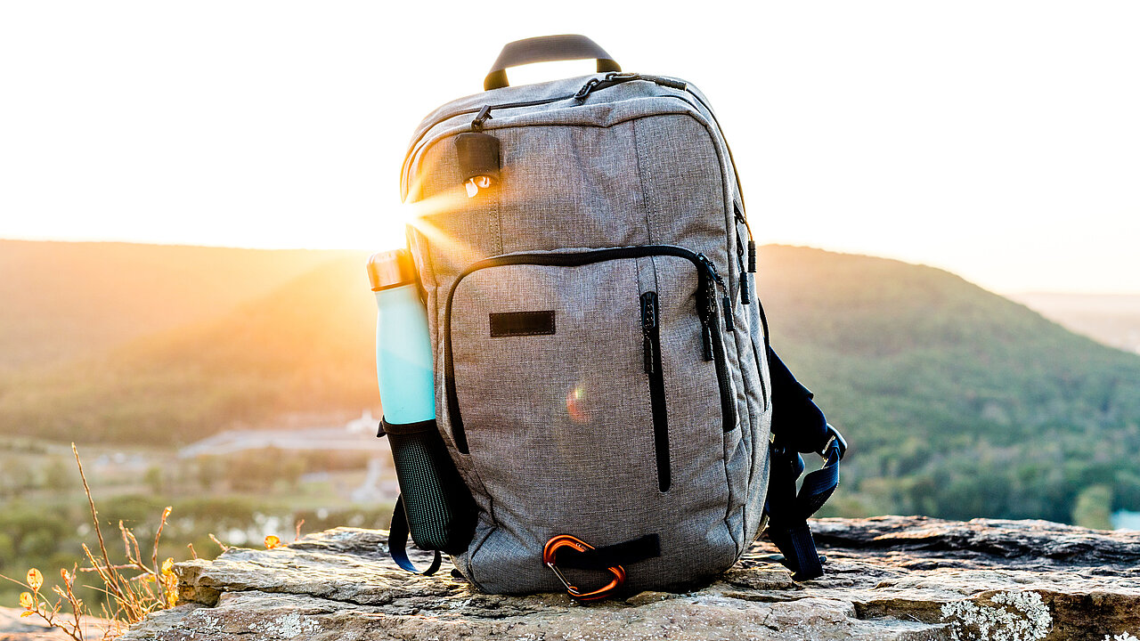 ein hellgrauer Rucksack auf Gras mit einem Sonnenuntergang im Hintergrund